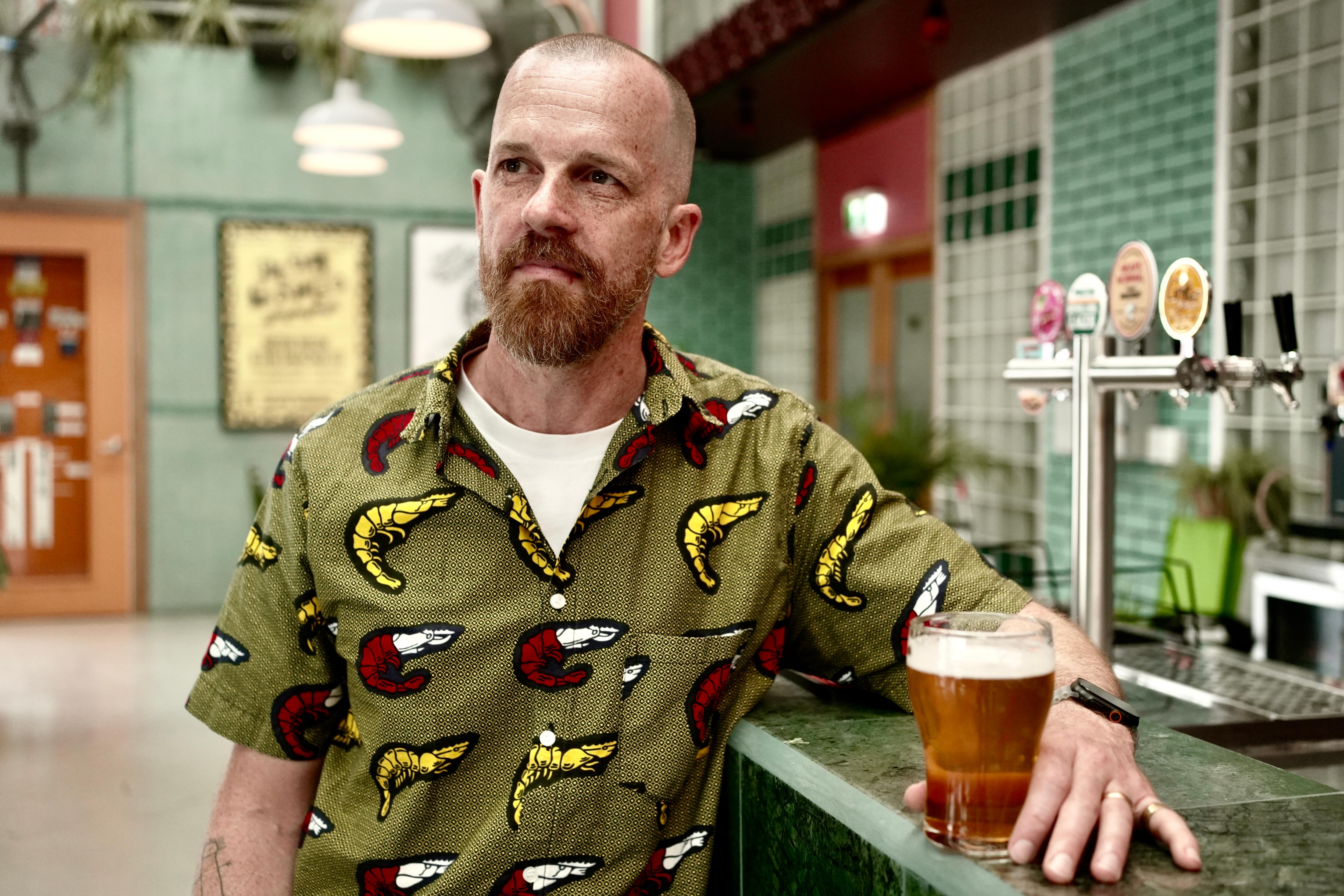 カラフルなシャツを着た男性がバーの前でノンアルコールビールを飲みながら立っている