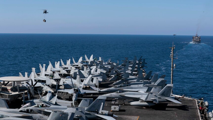 A bunch of fighter jets are seen parked on board the USS Abraham Lincoln aircraft carrier as ships are seen in the distance