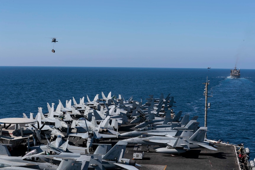 A bunch of fighter jets are seen parked on board the USS Abraham Lincoln aircraft carrier as ships are seen in the distance