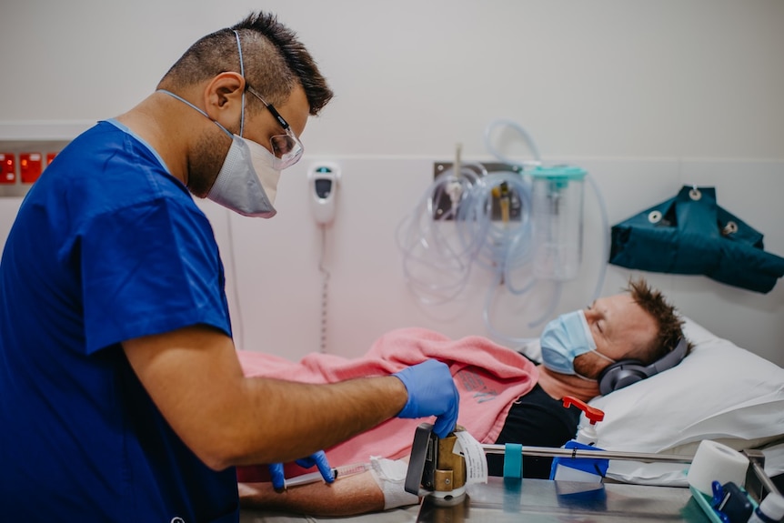 A patient lies in hospital bed receiving a syringe of fluid being injected into his arm.