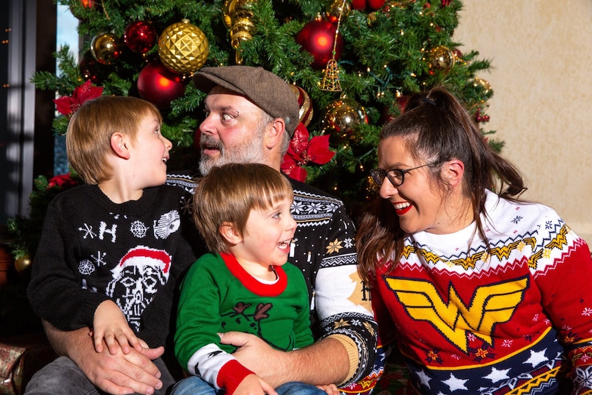 A family photo with a woman, man and two young boys wearing Christmas jumpers in front of a Christmas tree