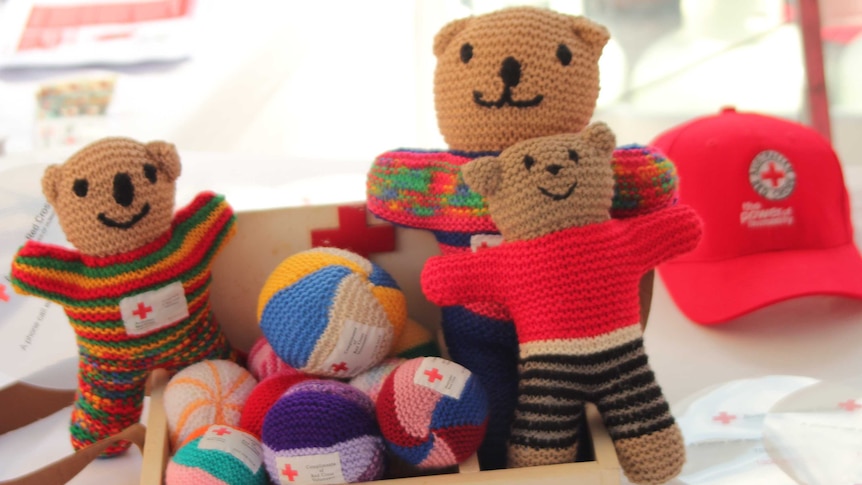 Three hand knitted, smiling Red Cross teddy bears sit in a box full of colourful knitted balls