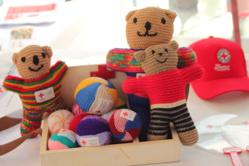 Three hand knitted, smiling Red Cross teddy bears sit in a box full of colourful knitted balls