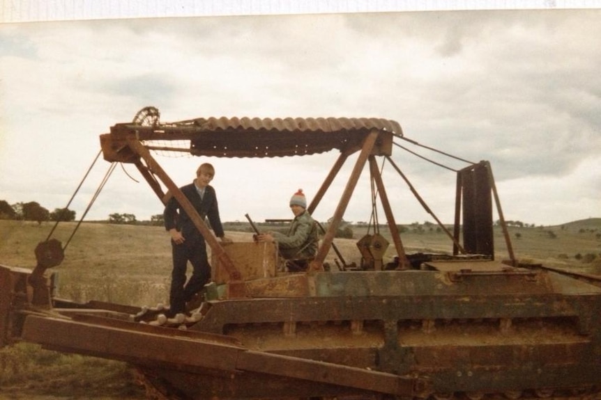 A faded colour photograph of two young men/boys on a tank equipped with a bulldozer blade and a shade of corrugated iron.