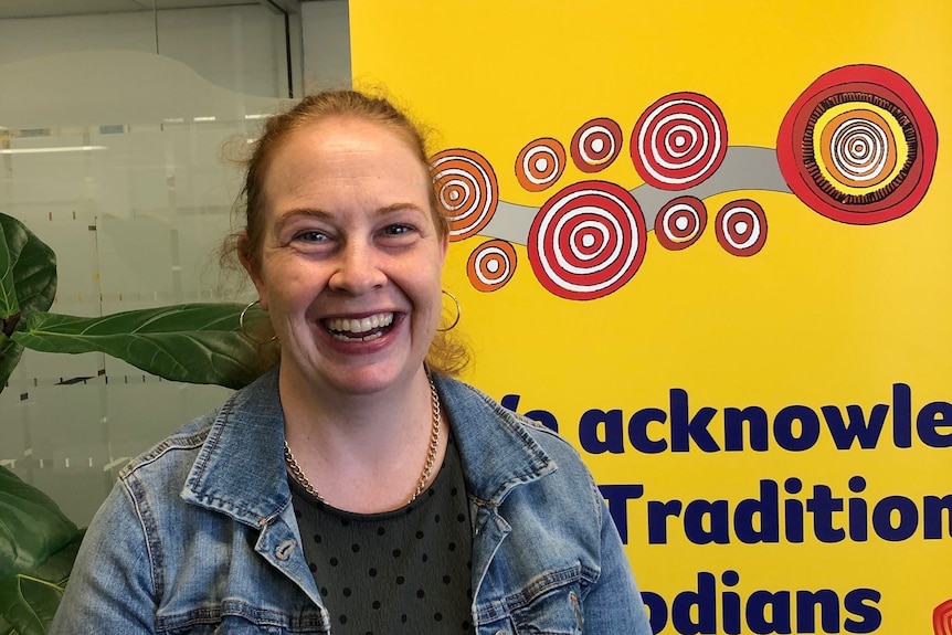 A smiling, curly-haired woman wearing a denim jacket stands in front of a brightly coloured sign.