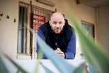 A bald man in his late 30s leans on a ledge looking at the camera with a plant in the foreground.