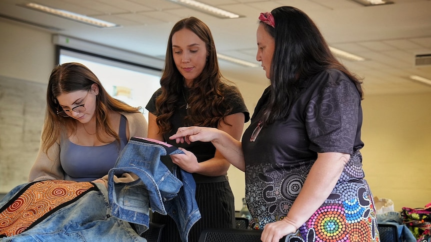 Three women behind a desk with chairs looking at denim pieces with Aboriginal art patched onto it