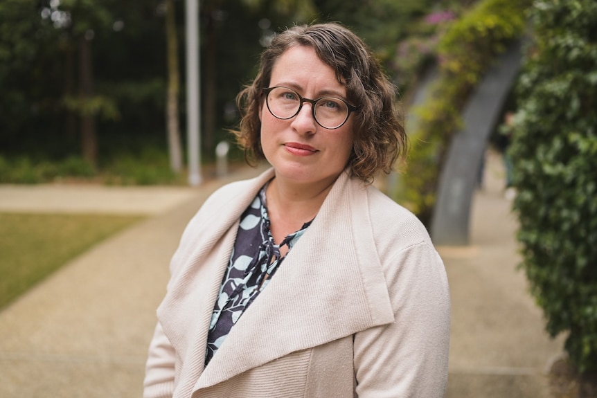 a woman wearing glasses and a white jacket. She has shoulder length hair. There are trees behind her.