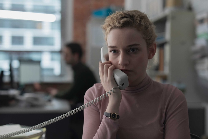 The actor Julia Garner in the film The Assistant, she's wearing a pink top, blonde hair, holding a landline phone up to her ear