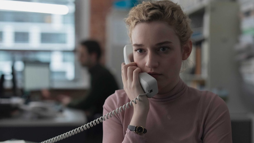 The actor Julia Garner in the film The Assistant, she's wearing a pink top, blonde hair, holding a landline phone up to her ear