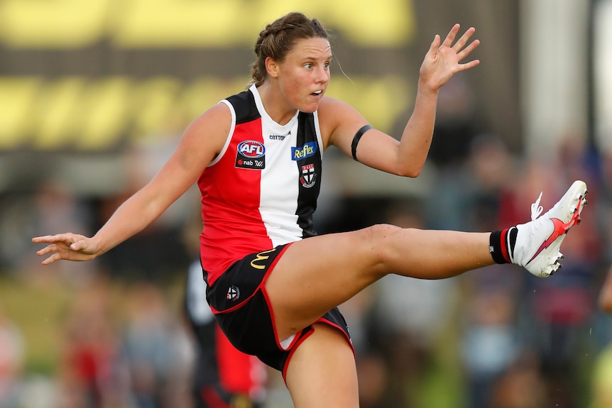 An AFLW player extends her leg after kicking the ball downfield during a game.
