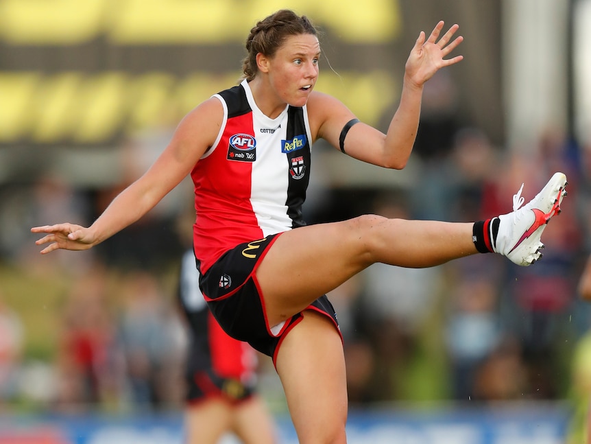 An AFLW player extends her leg after kicking the ball downfield during a game.