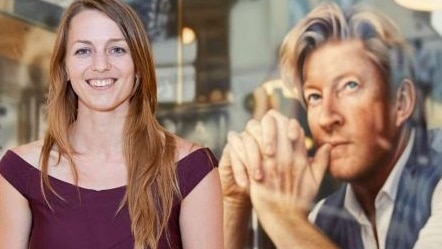 Smiling woman with long light brown hair wearing shortsleeve burgundy top & floral skirt, in front of portrait of David Wenham.