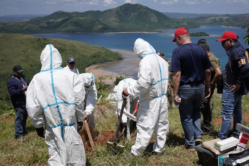 People wearing full protective white suits dig in the dirt on top of a grassy hilltop looking out to sea