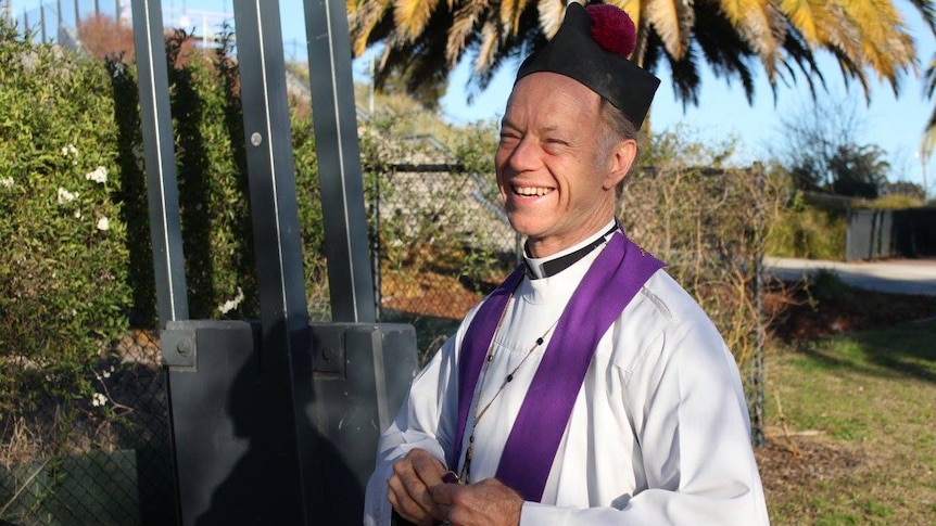 A priest in clerical robes and cap smiles in a backyard.