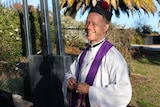 A priest in clerical robes and cap smiles in a backyard.