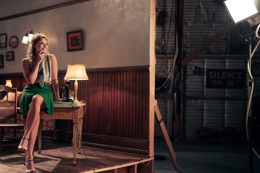 A blonde woman in her 30s, wearing a green skirt, smiles brightly, sitting with her legs crossed on a desk on a film set.