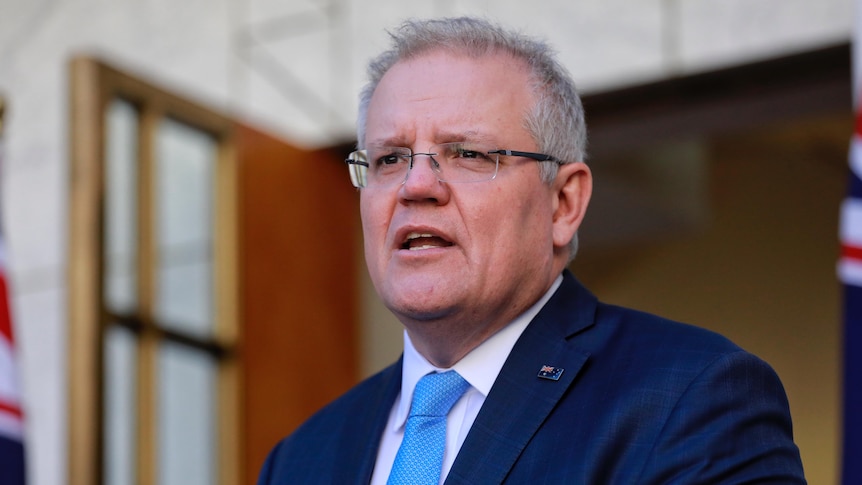 A man with grey hair and glasses wearing a navy suit and a light blue tie mid-sentence