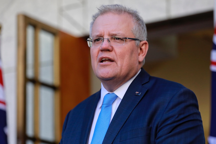 A man with grey hair and glasses wearing a navy suit and a light blue tie mid-sentence