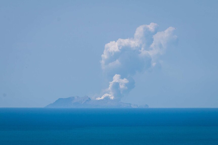From a distance over clear blue water, you see a plume of white smoke coming from Whakaari/White Island on a clear day.