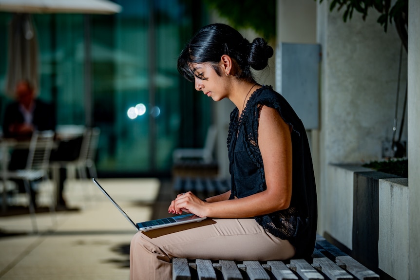 A woman in light pants, black top, black hair, sits on shady bench working on her laptop on her lap. 
