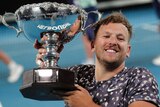 Dylan Alcott smiles while holding up a big silver trophy on the tennis court.