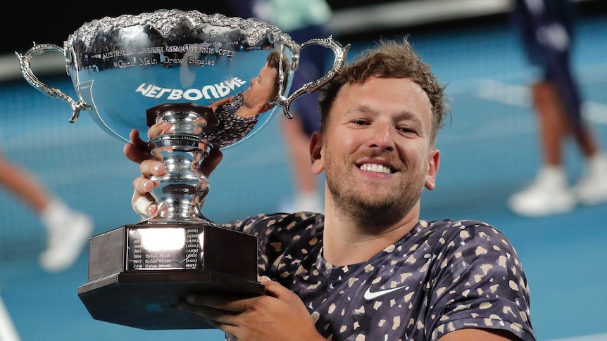 Dylan Alcott smiles while holding up a big silver trophy on the tennis court.