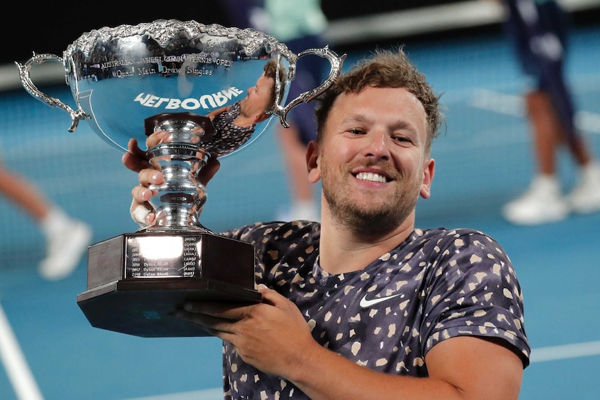 Dylan Alcott smiles while holding up a big silver trophy on the tennis court.