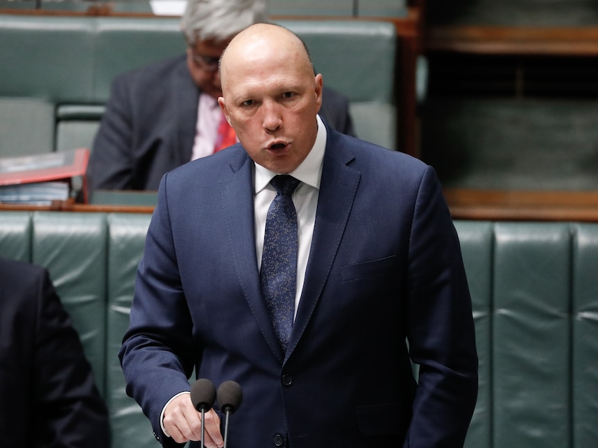 A bald man wearing a suit and tie mid-sentence standing at a lectern