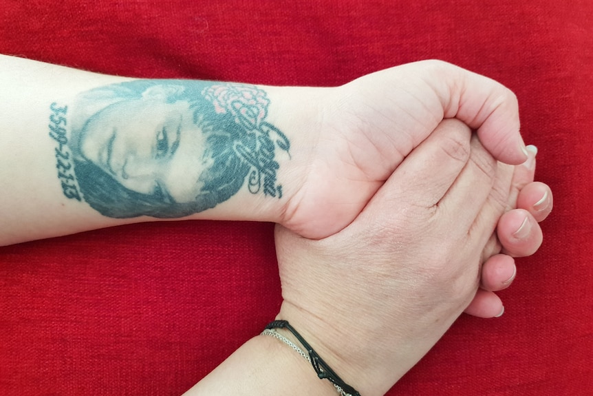 Two hands clasped together with the left wrist turned to show a tattoo of a young girl's face