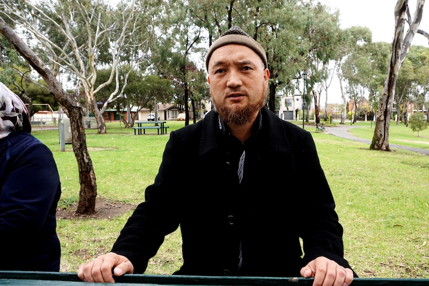 Man in black suit sits in park. Trees in background.
