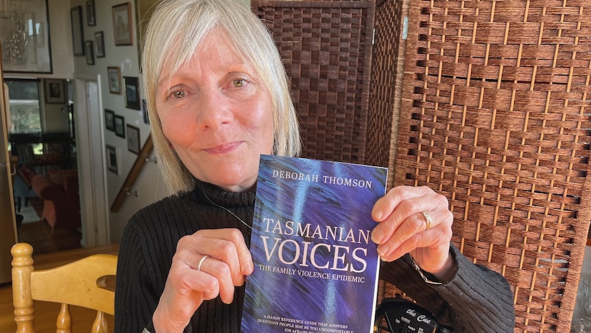 A woman with silver hair holds a book with the title Tasmanian Voices