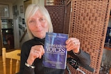 A woman with silver hair holds a book with the title Tasmanian Voices