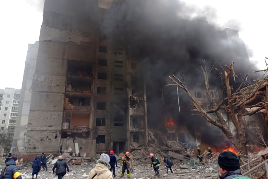 Smoke and flame are seen coming out of a multi-storey city building, firefighters below.