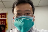 A man with a medical mask and glasses takes a selfie. He has short dark hair.