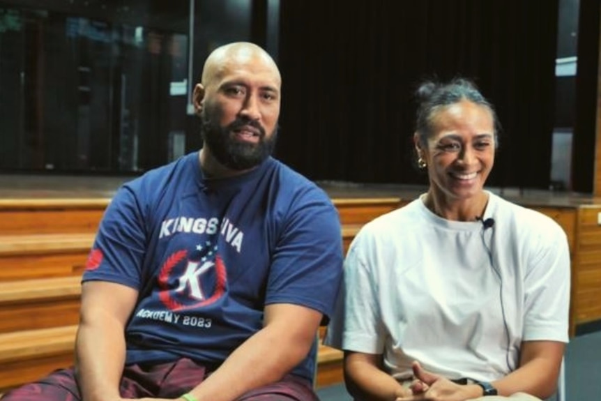 Dad Whiteny wears blue tee and sits next to his smiling wife Heather wearing white tee in classroom.  