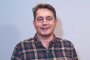 A head and shoulders image of a man in a checked shirt holding a glass of cider.