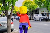 A man in a large Lego man suit waves in the middle of a tree-lined street.