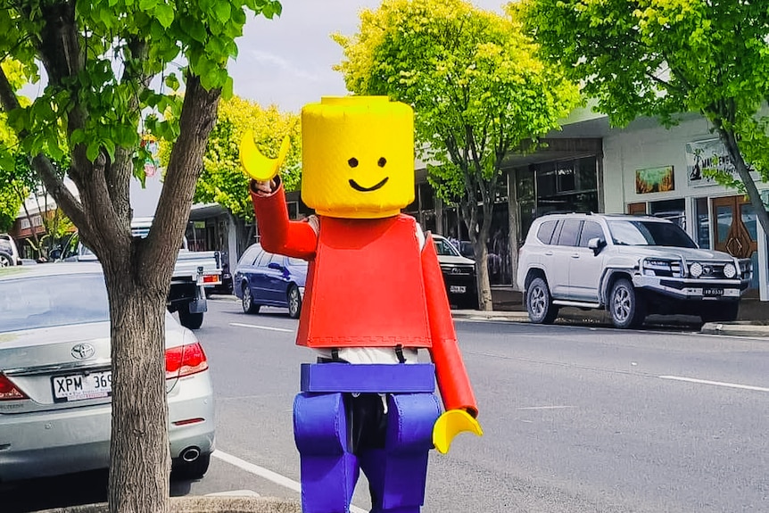 A man in a large Lego man suit waves in the middle of a tree-lined street.