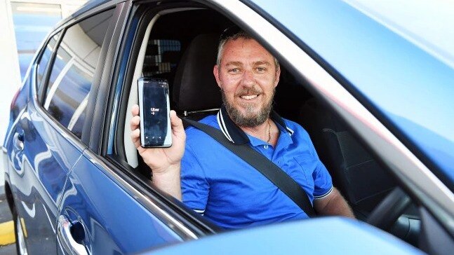 A man in a blue shirt sits in his car holding up his phone with the uber app on it