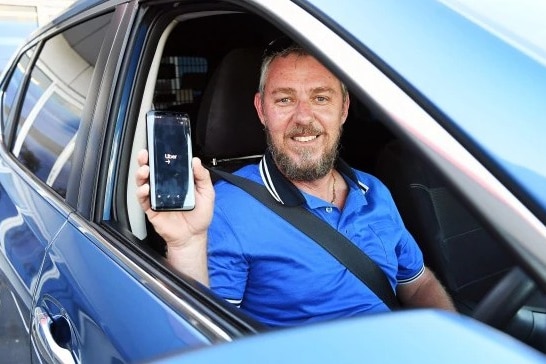 A man in a blue shirt sits in his car holding up his phone with the uber app on it