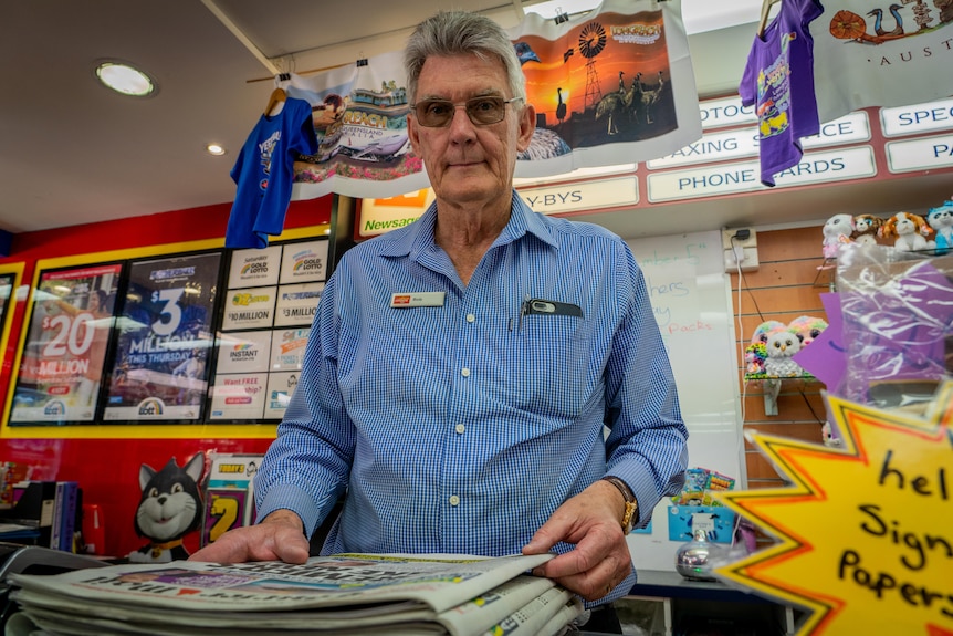 An older man stands behind a newsagents counter with a stack of newspapers in front of him. He is frowning.