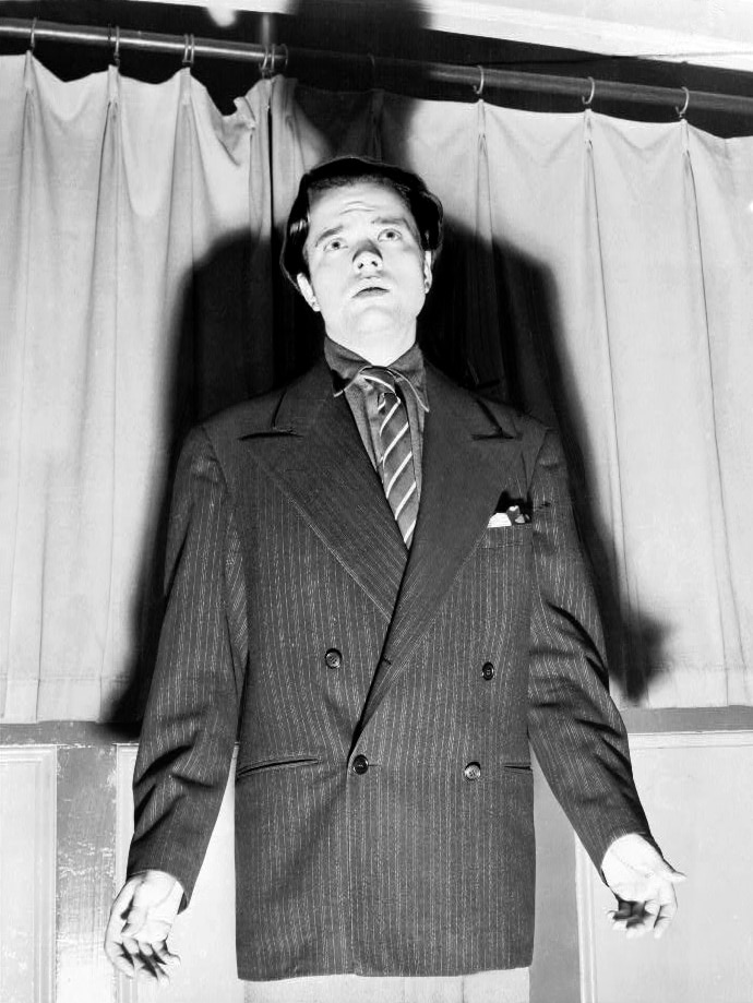 A man in a black and white photo wears a suit and tie with a long shadow behind him 