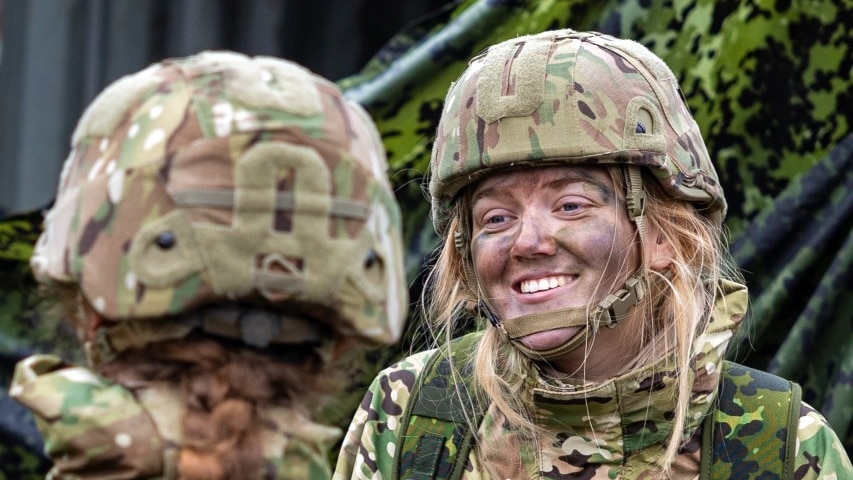 A young woman wearing a military uniform smiles at another person in uniform, who can only be seen from behind.