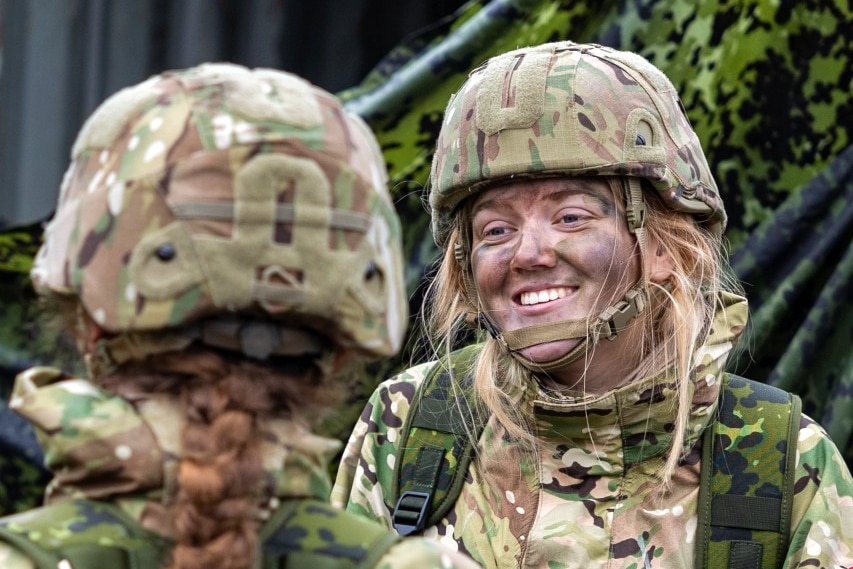 A young woman wearing a military uniform smiles at another person in uniform, who can only be seen from behind.