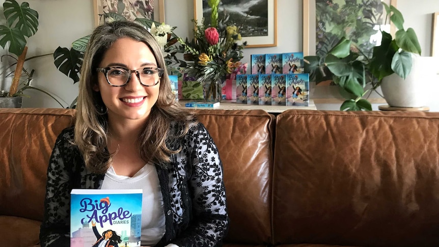 Smiling Latina woman in glasses sits on a couch holding a young readers' book called Big Apple Diaries
