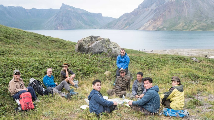 A group of men and women sit on the grass next to a river and mountain.