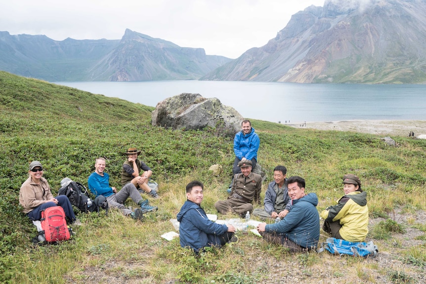 A group of men and women sit on the grass next to a river and mountain.