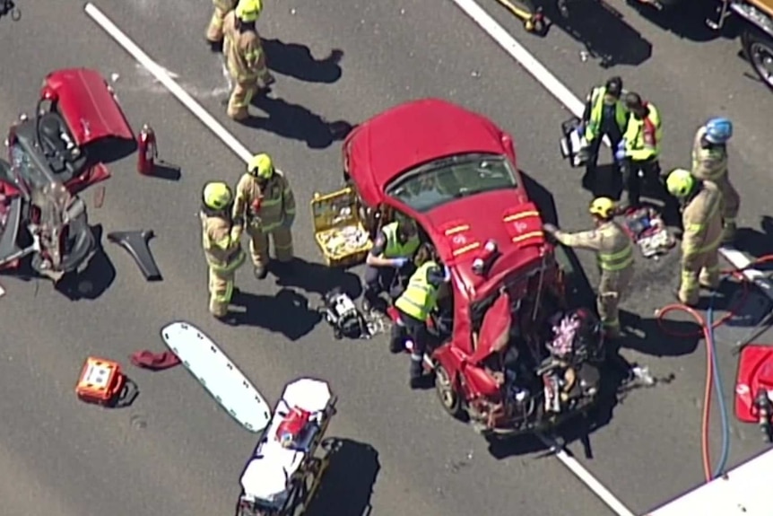 About 10 emergency workers wearing high-vis surrounded a smashed up red sedan, with a red car door lying meters away on the road