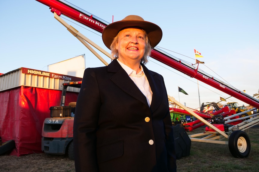 A lady with blonde hair and an akubra hat stands infront of a red crane at AgQuip.